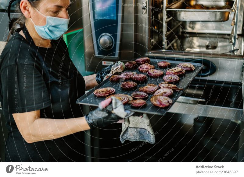 Crop cook in respirator with roasted onion on baking pan chef woman oven focus kitchen restaurant coronavirus ingredient food serious busy glove concentrate