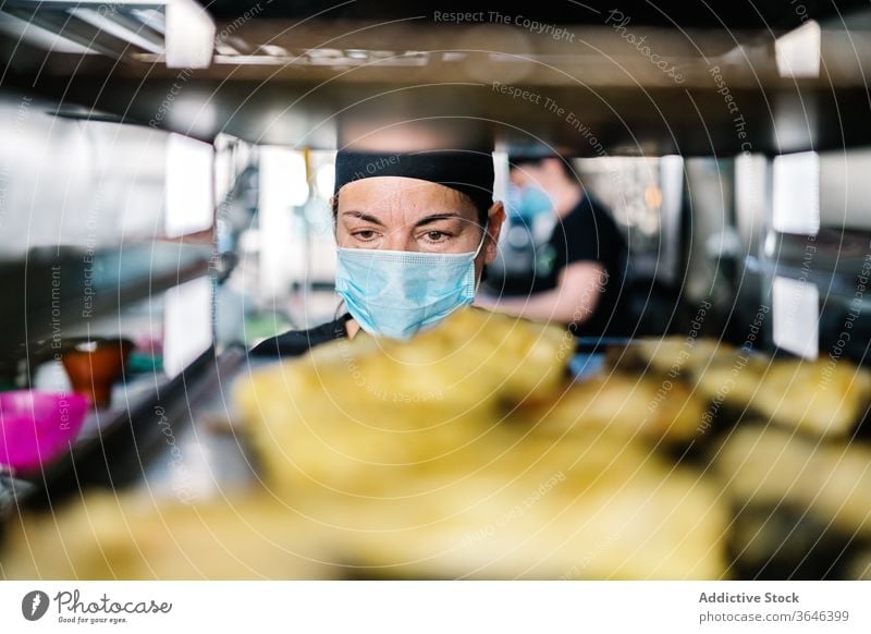 Female kitchen worker in face mask against pan rack woman chef food steel respirator coronavirus concentrate contemporary kitchenware colleague focus tasty busy