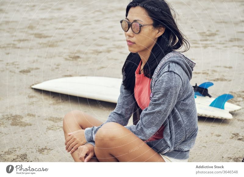 Calm Asian woman sitting on beach near surfboard surfer sandy rest thoughtful casual sunglasses seaside relax young sporty tranquil lifestyle nature seashore