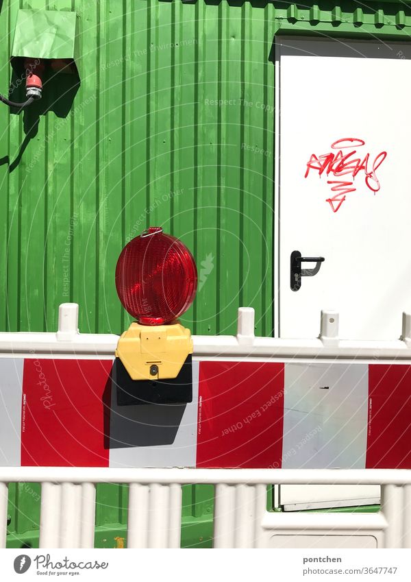 A red and white striped barrier with lamp in front of a building site with a green container, which is smeared with graffiti. Construction work. Protection against accidents