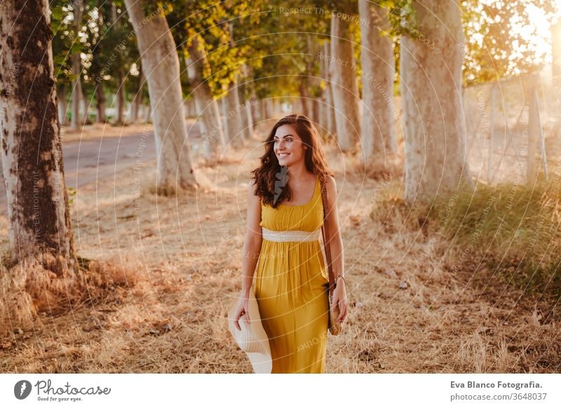 portrait of young beautiful woman wearing a yellow dress walking by a path of trees. Summertime and lifestyle adventure back beauty bright brunette casual