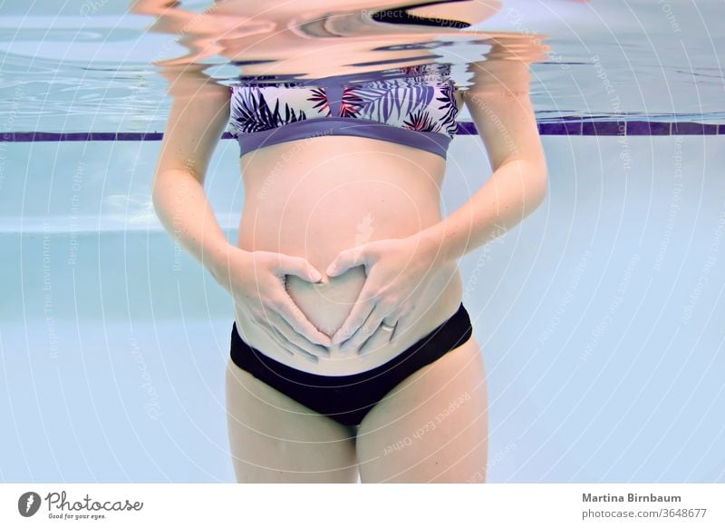 Pregnant woman with baby belly forming a heart with her hands, underwater capture motherhood pregnancy pregnant pool swimming bikini summer young beautiful blue