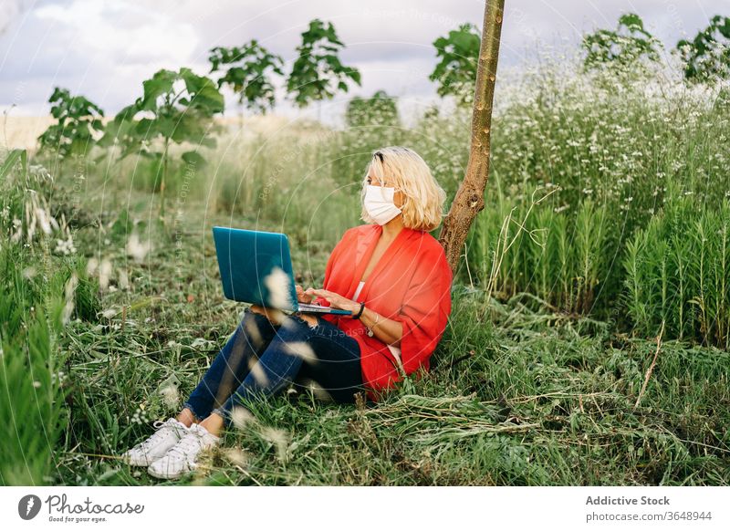 Focused woman in respirator working on laptop on grassy meadow using modern countryside nature coronavirus calm tree portable lawn casual mask peaceful rural