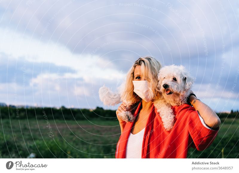 Content woman in respirator with cute little dog on shoulders bichon frise countryside nature funny coronavirus content casual pet puppy white covid 19 epidemic