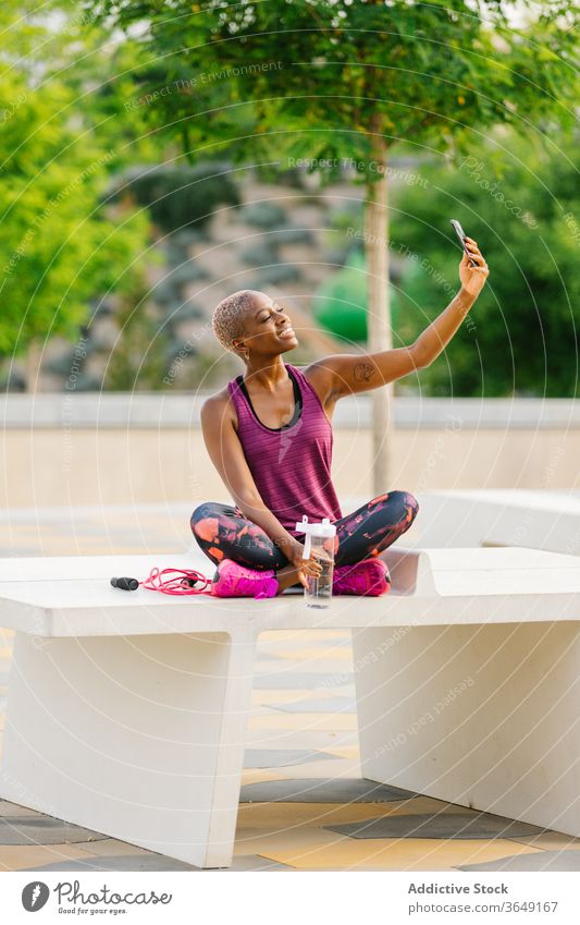 Cheerful black sportswoman taking selfie on smartphone after workout break summer city sportswear bench bottle using gadget legs crossed device cellphone tree