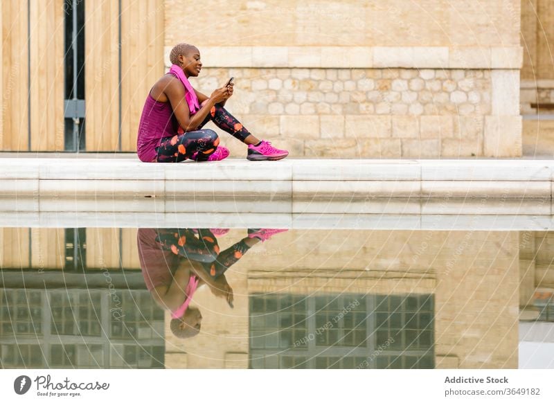Black female athlete surfing internet on smartphone near city fountain sportswoman social media break towel reflection embankment using gadget device cellphone
