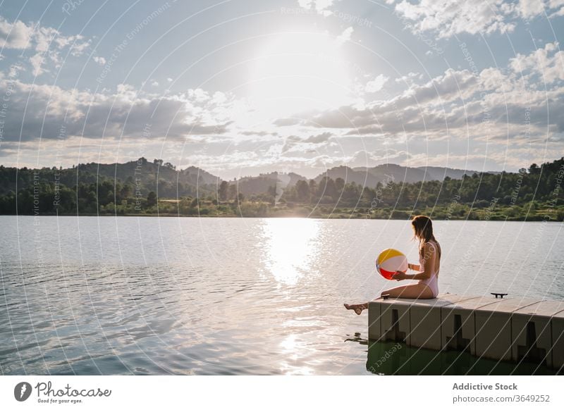 Woman sitting in edge of in pier near lake woman beach ball play pond summer swimwear holiday vacation enjoy inflatable rubber water relax nature playful