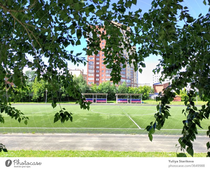 Empty green soccer field near a school and the apartment house in Russia. Stadium and football field with empty spectator seats. Keeping fit and exercising outdoor. Urban view in a frame of leaves.