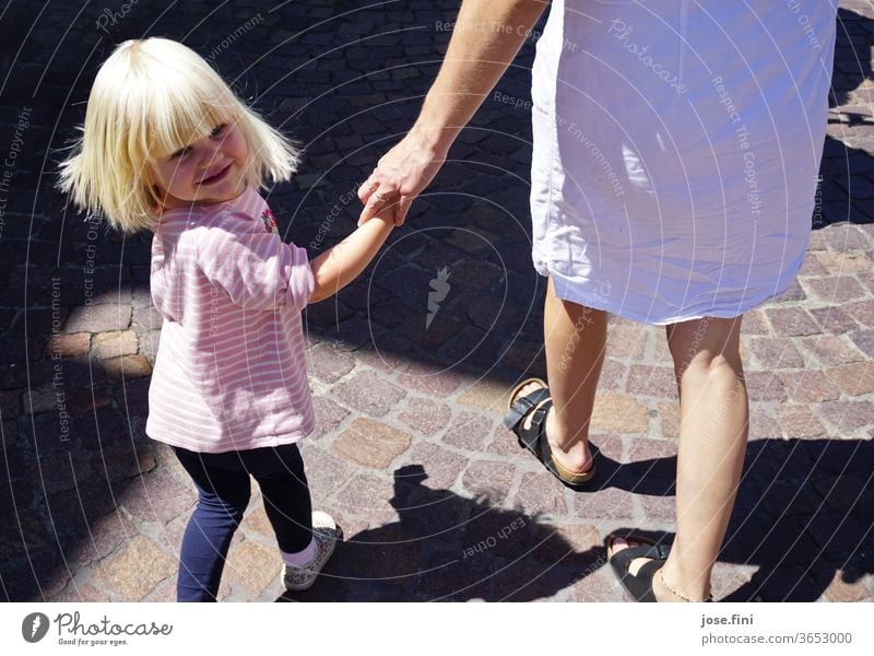 Little girl walks by the hand of her mother and looks back smiling with a swing in her hair. Child Parent with child Mother Walking To go for a walk smile Joy