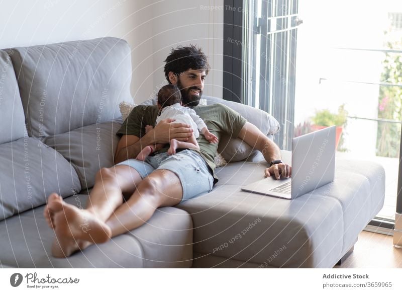 Busy father with infant using laptop baby work freelance sofa busy newborn project male fatherhood netbook home gadget remote browsing internet surfing online