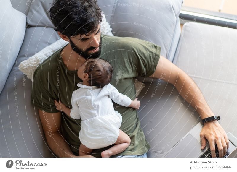Busy father with infant using laptop baby work freelance sofa busy newborn project male fatherhood netbook home gadget remote browsing internet surfing online