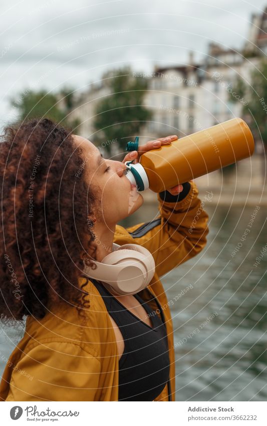 Thirsty ethnic sportswoman drinking water after working out near river thirst bottle headset sportswear break tree using gadget headphones device urban plastic