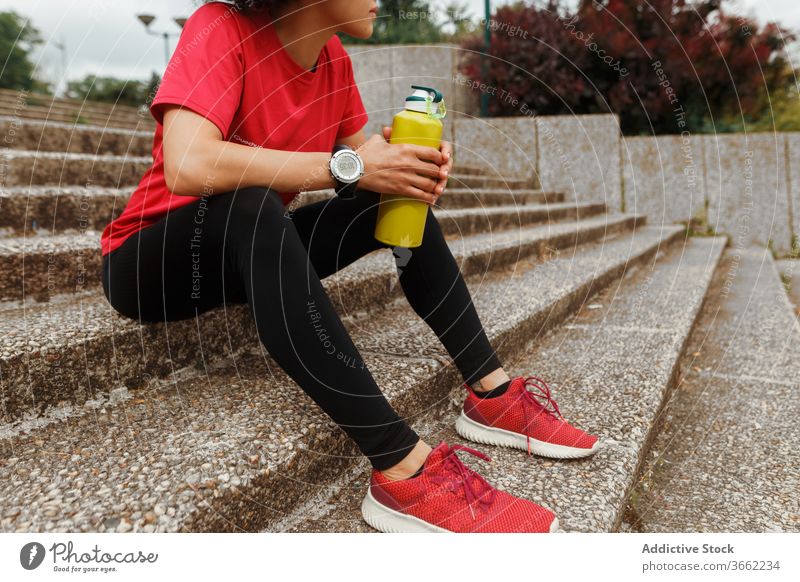Thirsty ethnic sportswoman drinking water on stairs after workout thirst thermos sportswear break hydrate eyes closed relax rest staircase athlete device urban