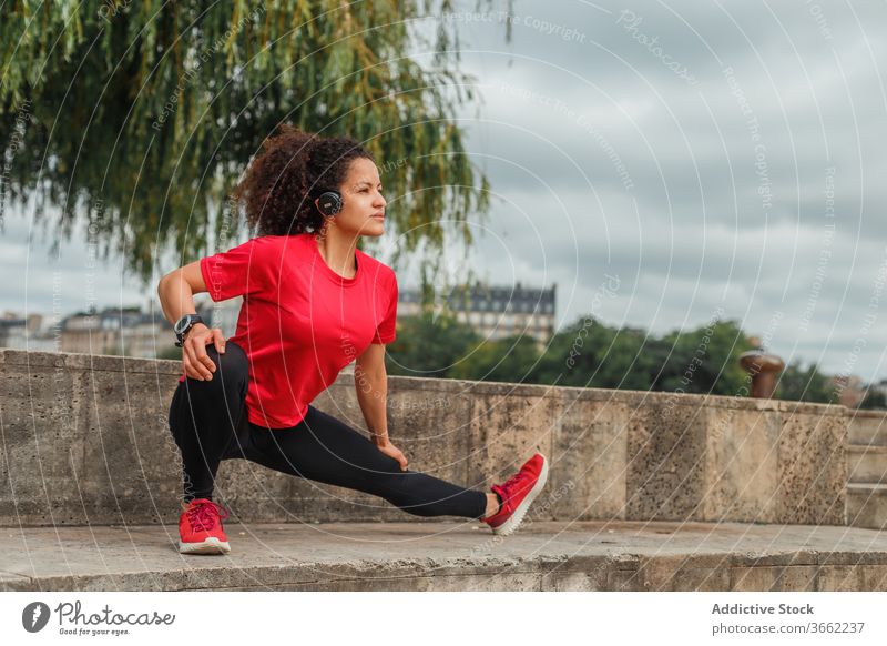 Ethnic sportswoman in headphones stretching leg while warming up outdoors squat warm up workout exercise wireless listen music headset flexible using device
