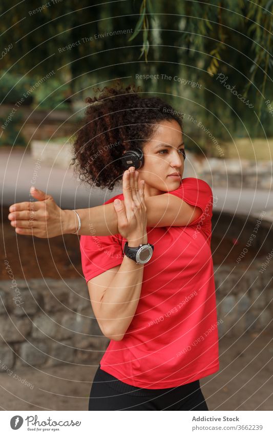Ethnic sportswoman in headset stretching arm during workout in park athlete smart watch exercise listen music wireless reached city warm up using device gadget