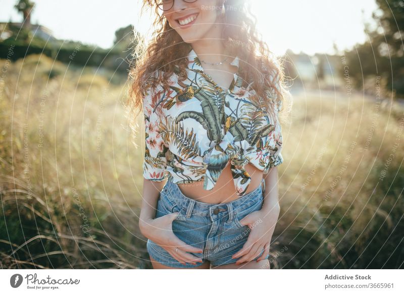 Crop glad woman in eyeglasses standing in field in summer hand on hip happy beauty grass lifestyle idyllic feminine summertime toothy smile harmony faded