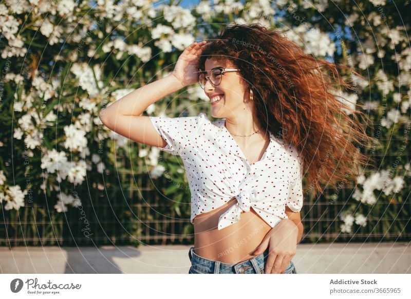 Positive female with wavy hair standing near blooming flowers woman touch hair fence happy beauty lifestyle belly idyllic feminine summertime toothy smile