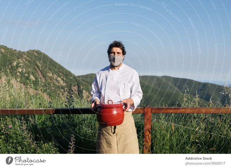 Chef in uniform with saucepan in mountainous valley man cook countryside picturesque nature soup dinner dish meal prepare male apron mask landscape professional