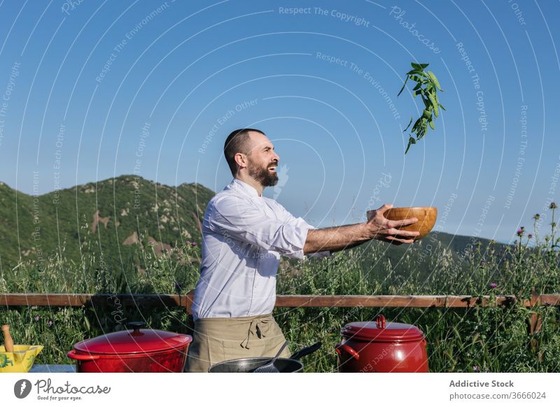 Positive male chef throwing veggies during cooking preparation outdoors man lunch nature toss vegetable bowl healthy happy food saucepan joy beard apron fresh