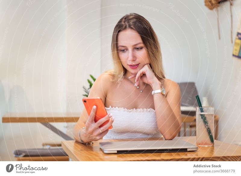 Positive young woman using smartphone while sitting at table with laptop and juice read message break cafe touch hair pensive device gadget female summer outfit