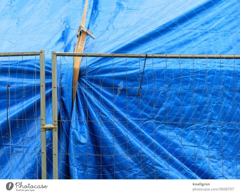 Large blue tarpaulin secured with a lashing strap behind a metal construction fence Protection Safety Covers (Construction) Structures and shapes