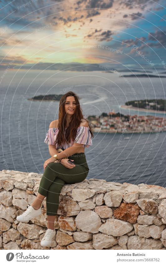 Attractive brunette sitting on a stone wall on a warm summer windy day. Fashionable girl posing with the adriatic sea and the small town of primosten in the background. Beautiful golden sunset