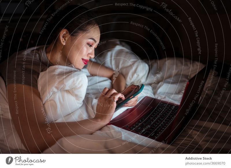 Positive Asian woman lying in bed with laptop and smartphone relax internet comfort domestic cheerful night using device gadget netbook enjoy smile glad
