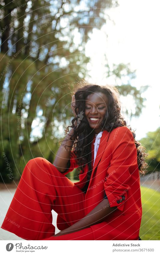 Cheerful black model in red outfit sitting on bench outdoors fashion appearance colorful cloth glamour elegant cheerful park woman vogue suit apparel slim
