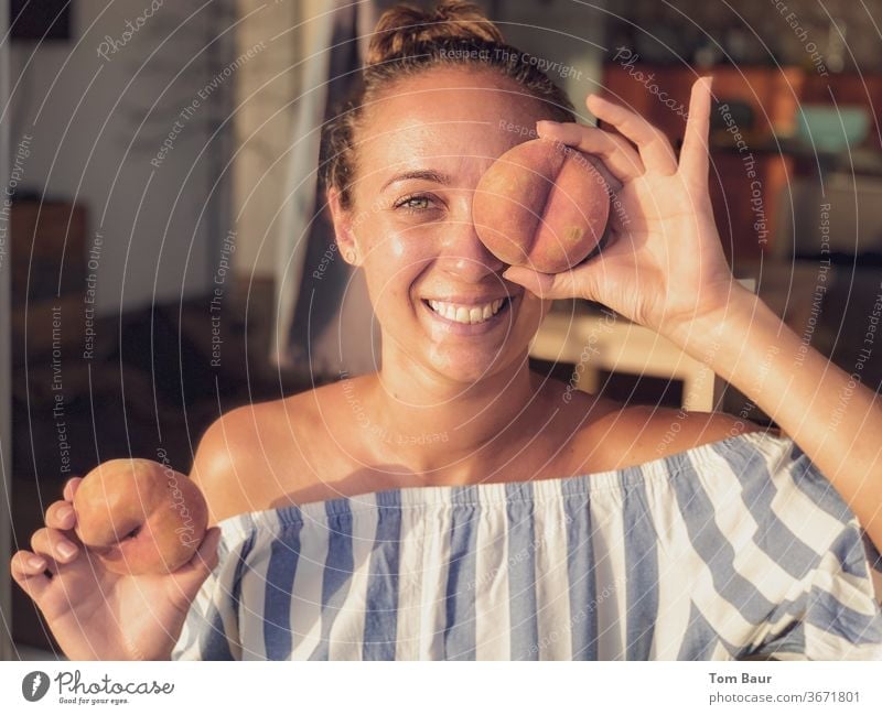 Young woman with brunette hair holds a peach in front of her eye Woman Peach Eyes Laughter happy fortunate Comical fun blue white portrait Shoulder