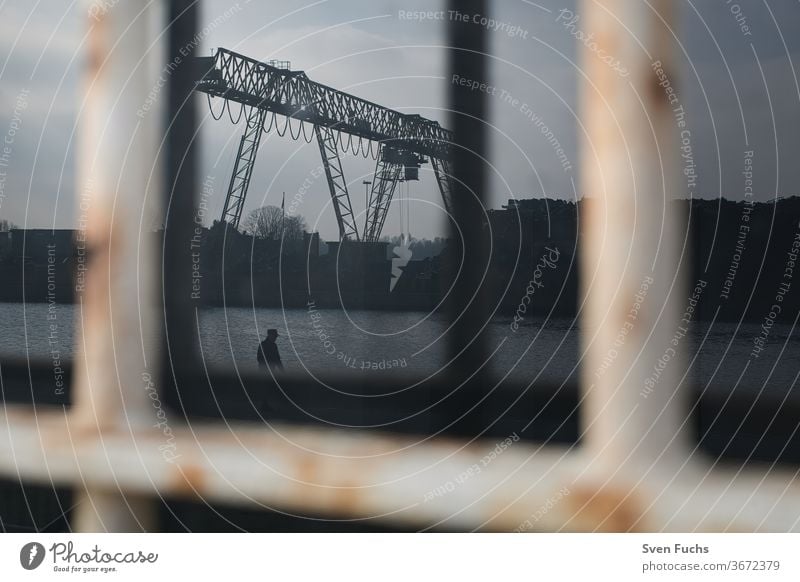An industrial crane is reflected in a dirty window. A single man walks past the quay Industrial Crane Window pane reflection frowzy Industry Landscape built
