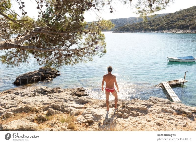 Rear view of man wearing red speedos tanning and realaxing on wild cove of Adriatic sea on a beach in shade of pine tree. Relaxed healthy lifestyle concept.
