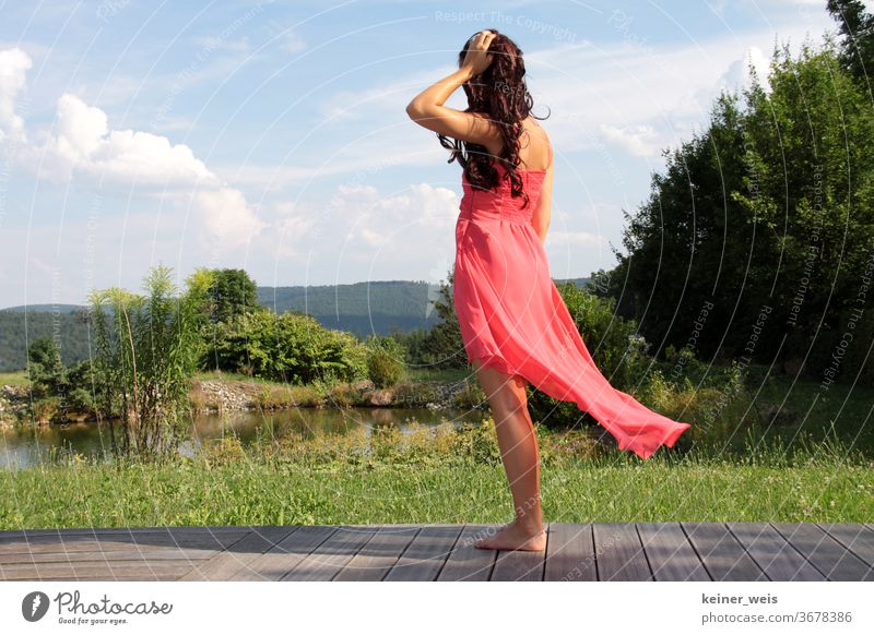 Young woman in flowing red dress stands barefoot on terrace in front of green landscape with small lake Woman Long-haired Dress Red Terrace Landscape Curl Lake
