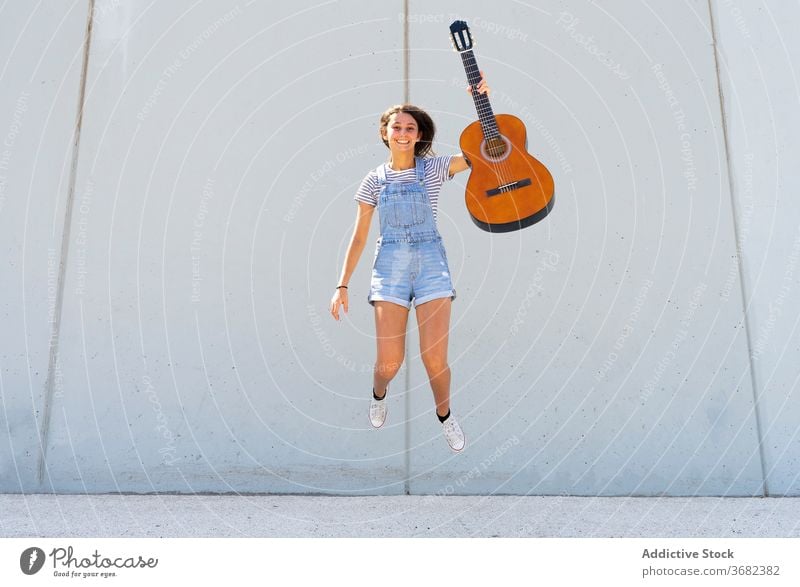 Optimistic teenager jumping with guitar musician smile wall building casual summer energy hobby girl happy acoustic modern city street cheerful urban joy