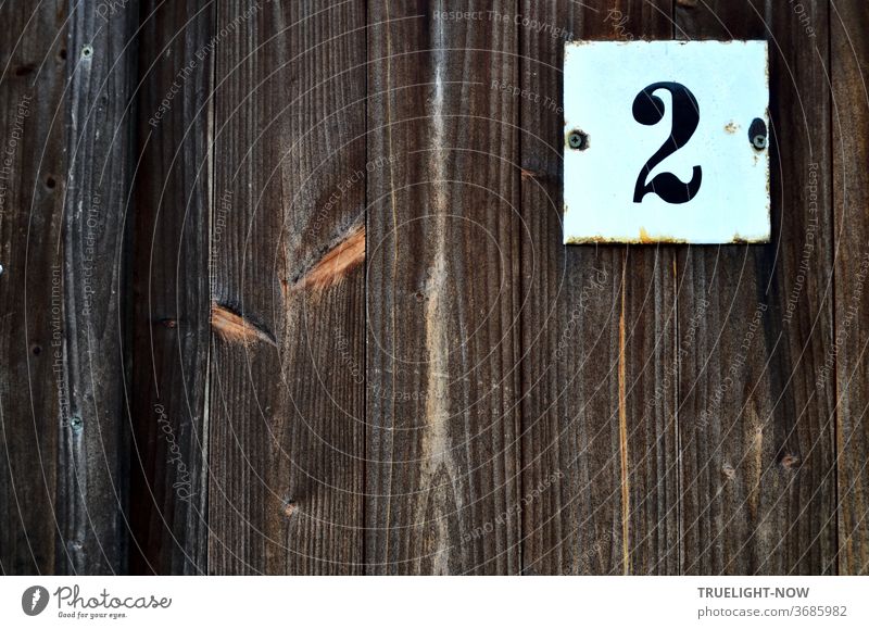 A white square metal plate is screwed to an old, dense fence made of wooden boards in vertical arrangement, on which a black, conspicuous number 2 in Antiqua script probably marks the house number