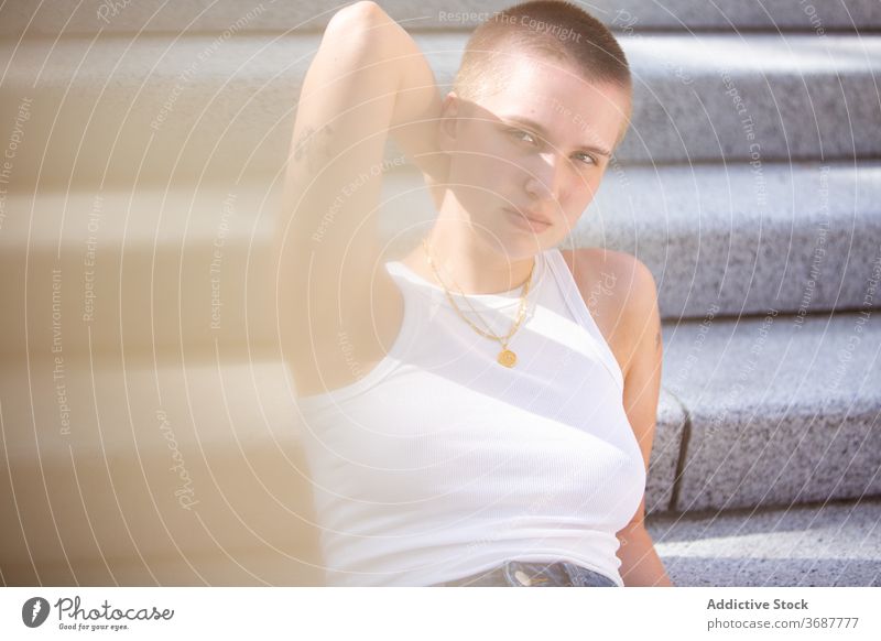 Young woman with shaved head resting on steps street city appearance subculture casual touch neck female young stair relax style trendy short hair serious