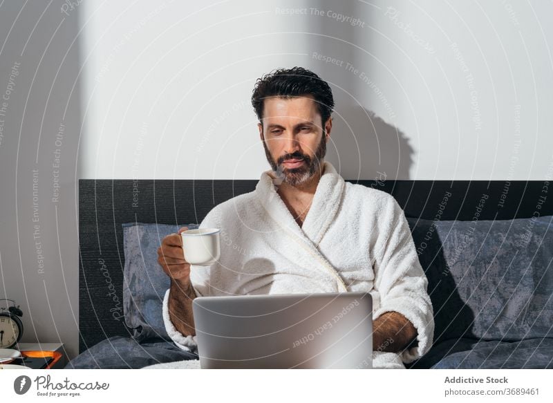 Calm man having breakfast on bed freelance coffee laptop work remote bathrobe using male businessman cup fresh morning drink device internet comfort online