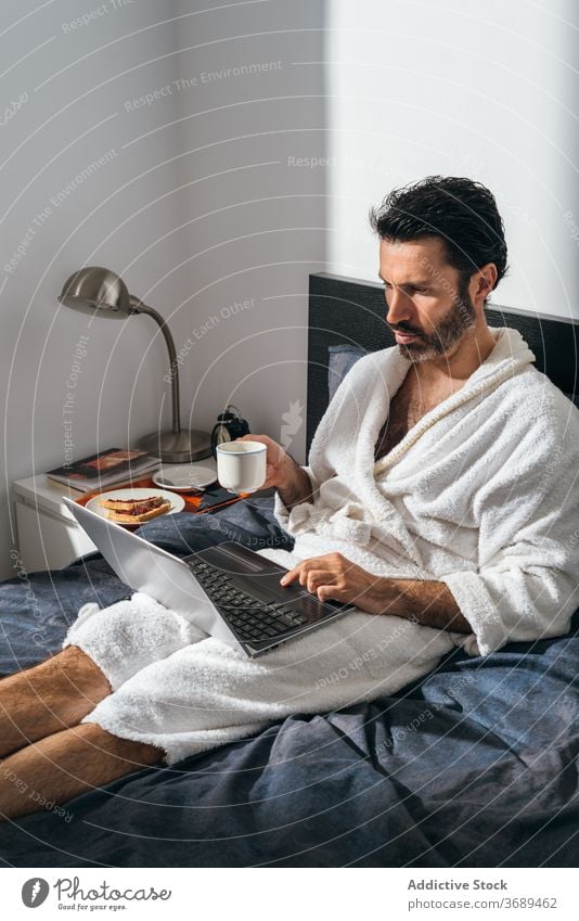 Calm man having breakfast on bed freelance coffee laptop work remote bathrobe using male businessman cup fresh morning drink device internet comfort online