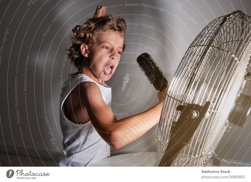 Child with arms raised holding a hairbrush that mimics a microphone singing in front of a fan indoors expressive proud play game joke fun emotion loud