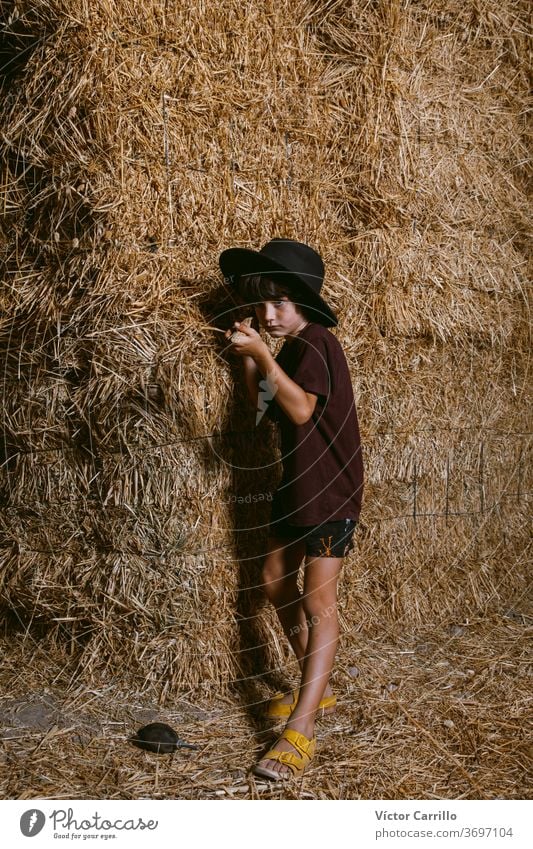 A young boy with a hat playing in the fields of the country side in summer in a straw bale background farmer boy crouching plant autumn kid cute fall plantation