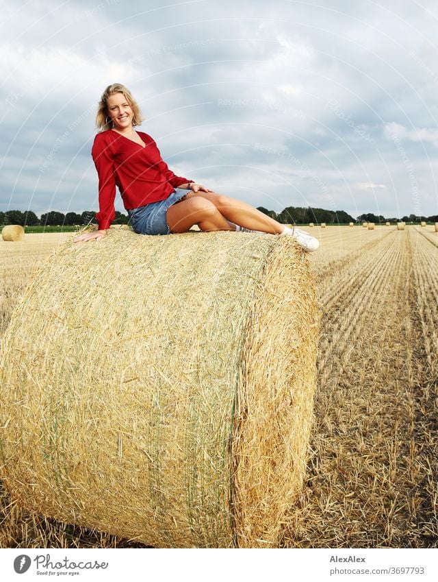 Portrait of a young woman on a bale of straw in the evening light Young woman Woman Blonde smile Red portrait Jewellery already Long-haired Landscape Clouds Sky