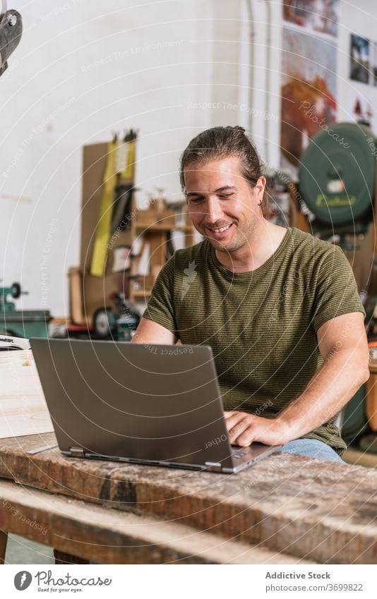 Smiling male woodworker using laptop in workshop carpenter man project workbench content netbook modern device busy workplace occupation professional