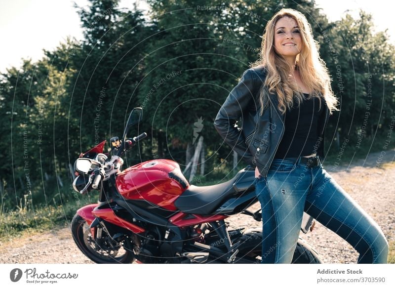 Cheerful biker near motorcycle in countryside motorcyclist modern sport motorbike woman transport cheerful leather jacket female trendy smile happy stand