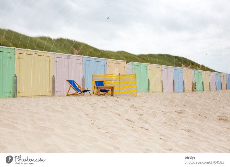 pastel-coloured beach huts in a long row. 2 empty deckchairs and a windbreak in front Beach Sand Pastel tone variegated Multicoloured wind deflector