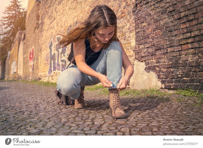 young woman in summer clothes ties her shoes crouching street outdoor beautiful concentrated bag wall brick Berlin lifestyle pavement teen teenager girl casual