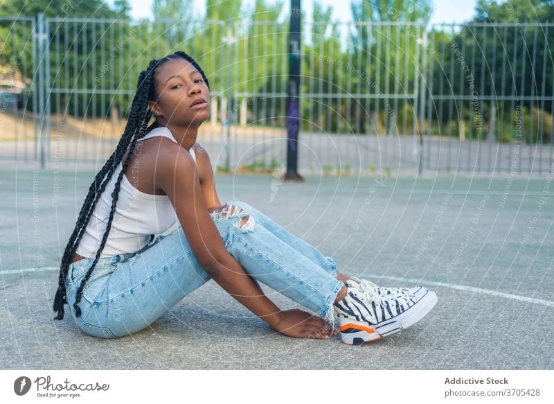 Young black woman sitting on sports ground urban style street style trendy hipster young braid ethnic millennial female jeans basketball outfit fashion casual