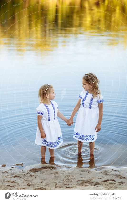 Two adorable little sisters laughing and hugging on warm and sunny summer day. two sisters in white dresses near the water. child family girl friend kid enjoy
