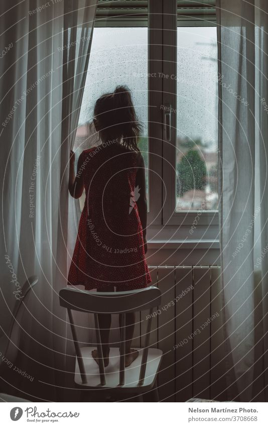 Little girl looking through the window in her bedroom in a rainy day. She is standing in a white chair, backward. We can see drops in the crystal. red child