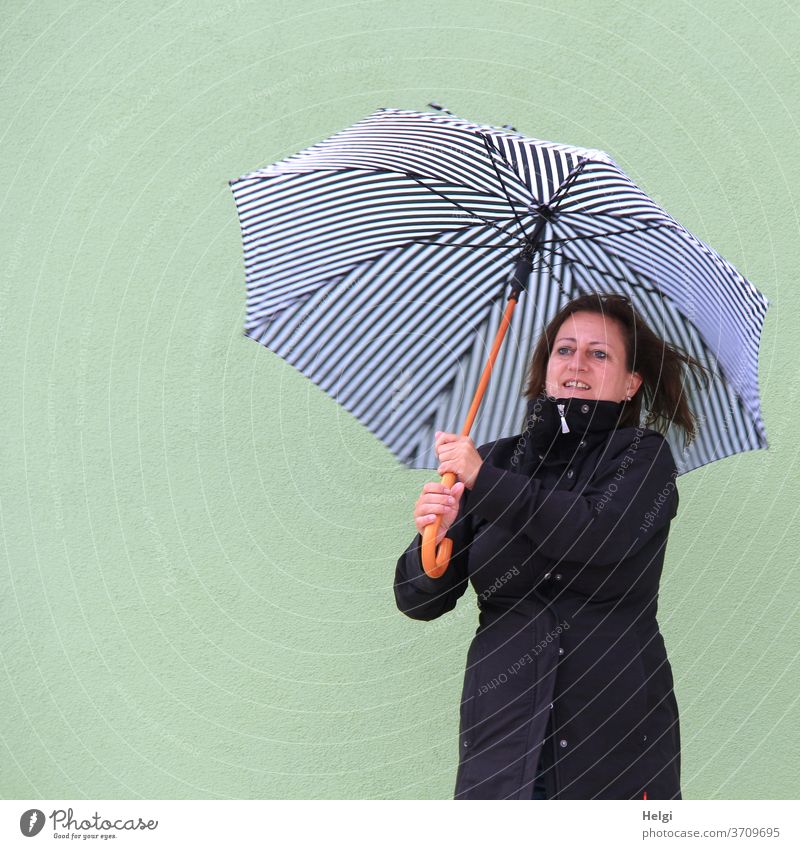 smiling woman in a black coat stands in front of a light green wall and holds a blue and white striped umbrella Woman Human being feminine Feminine