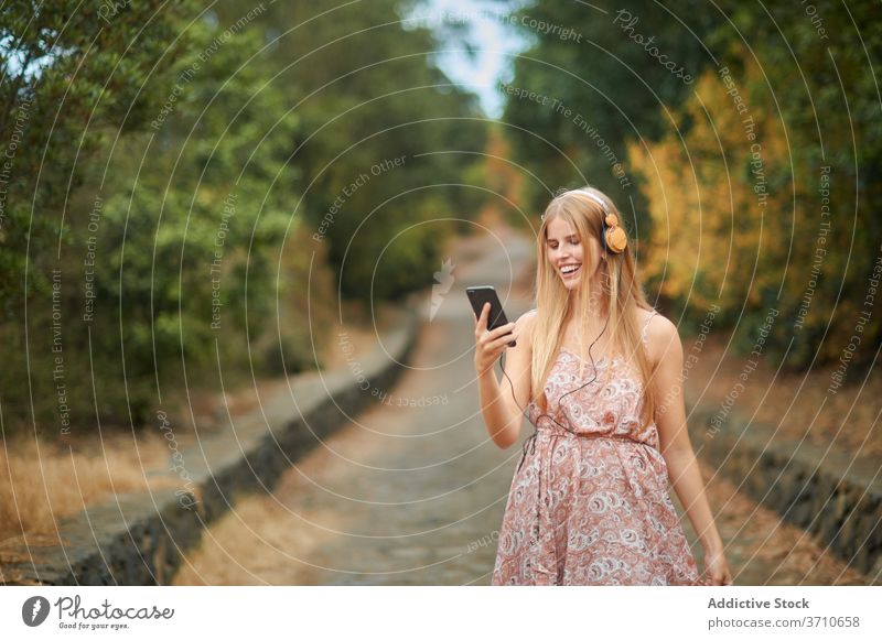 Smiling woman with smartphone and headphones walking in park happy listen using music summer enjoy young female browsing alley path device gadget lifestyle