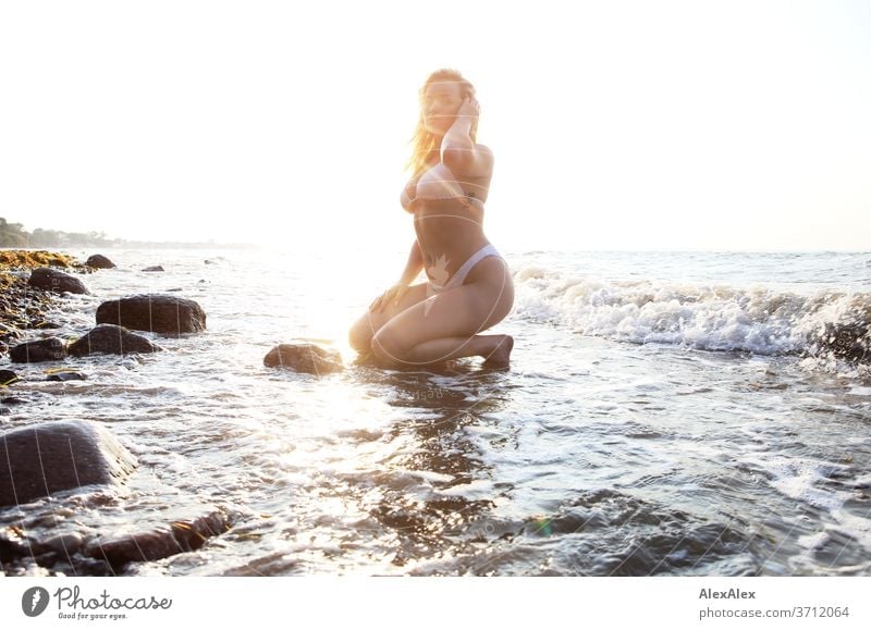 Portrait of a young blonde woman in a bikini, kneeling in the Baltic Sea surf Young woman Woman girl 18-20 years Blonde Slim already Curly sensual natural